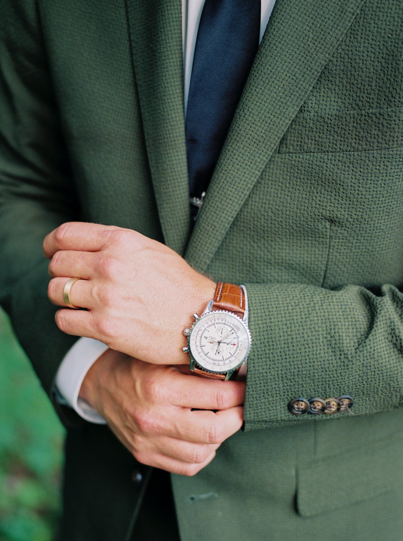  Closeup of groom’s adjusting brown leather wristwatch, wearing forest green Indochino suit and navy blue tie 