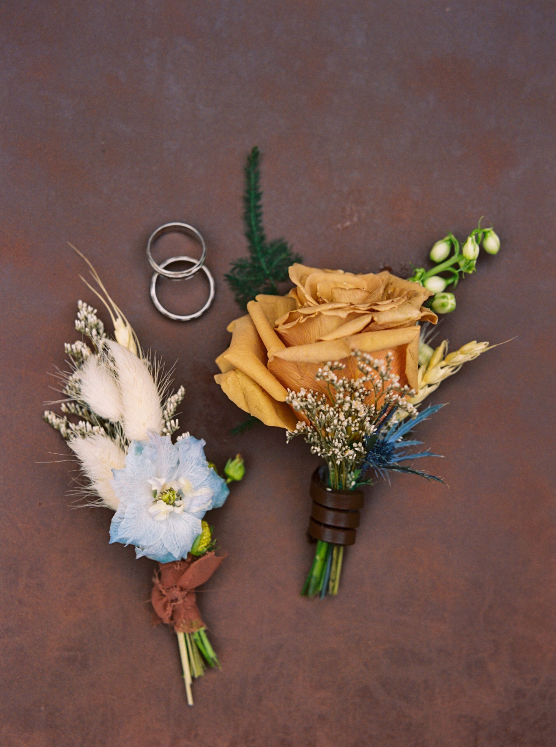  Boutonnieres by Little Thistle Floral Design and wedding rings on brown leather surface 