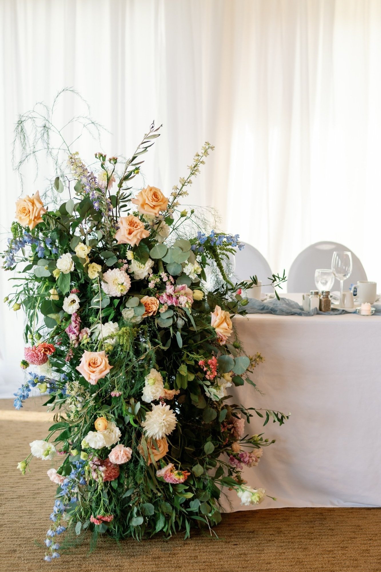 Floral pillar with peach roses, white dahlias, and blue delphinium next to a wedding head table