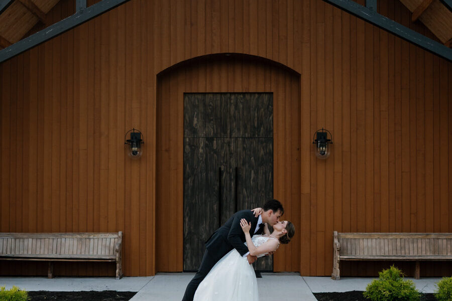 A groom dips his bride in front of a modern wood-clad building, both dressed formally, offering rustic winter wedding inspiration with warm architectural tones.
