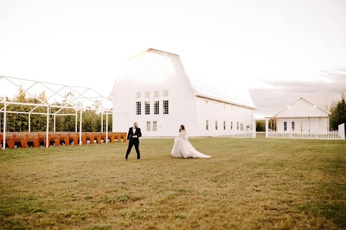A bride and groom dance in front of a large white barn at sunset, the wide lawn and soft golden light creating romantic countryside winter wedding inspiration.