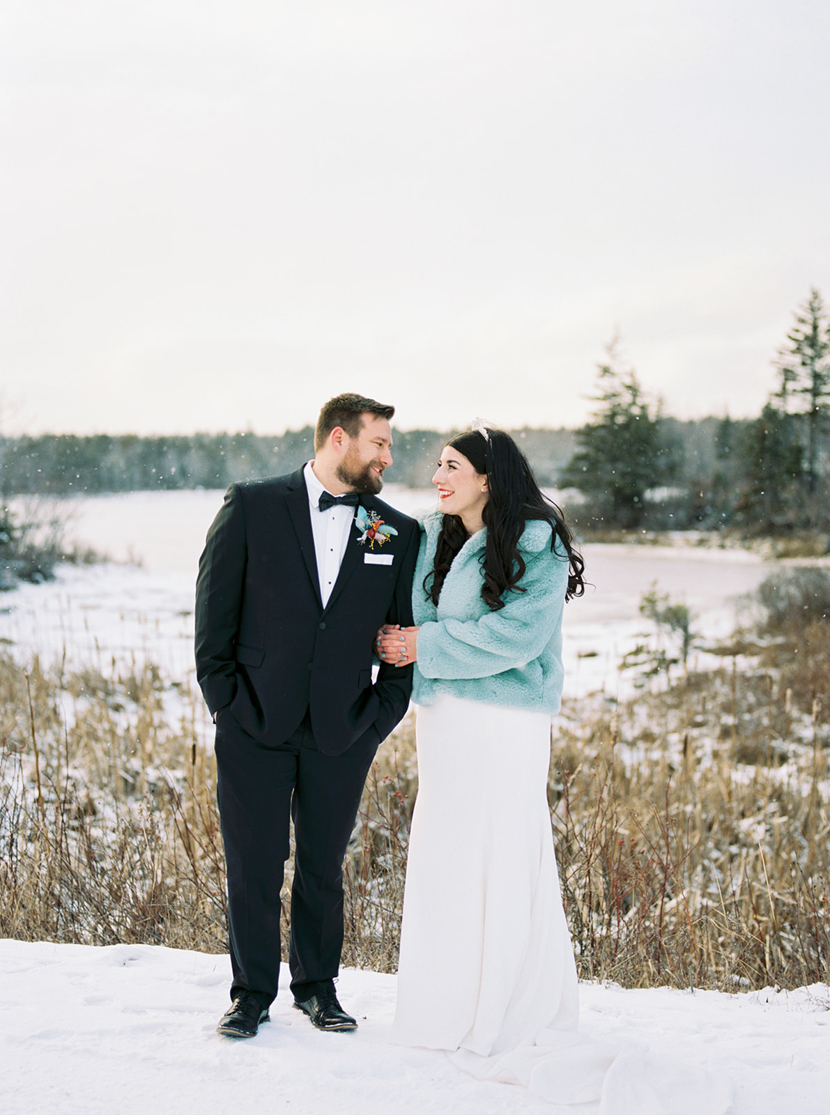 A bride and groom stand together in a snowy landscape, the bride in a teal faux-fur jacket and white gown and the groom in a tuxedo, sharing a warm moment for cozy winter wedding inspiration.