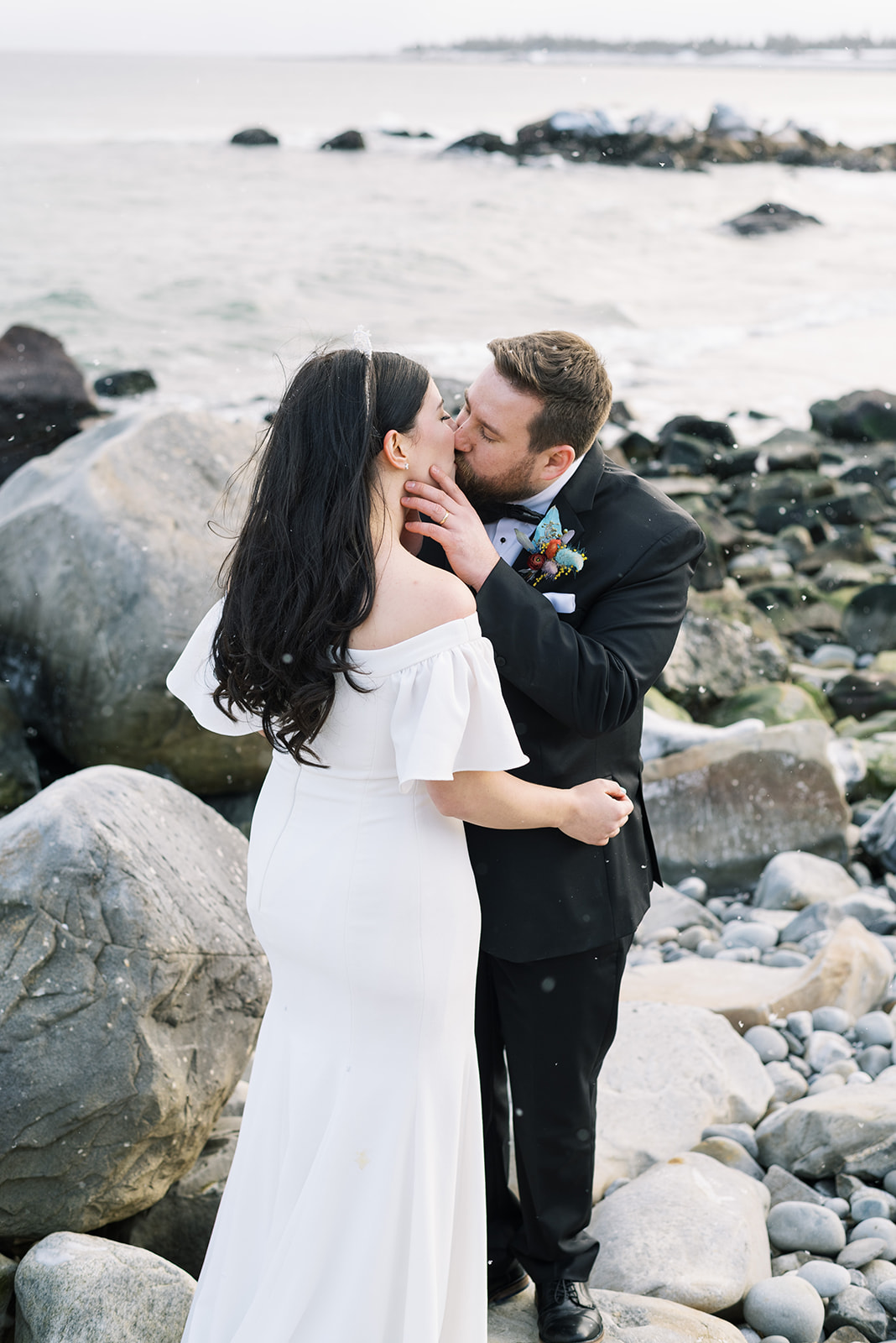 A bride and groom share a kiss on a rocky shoreline with snowflakes falling, the bride in an off-the-shoulder white gown and the groom in a black tuxedo