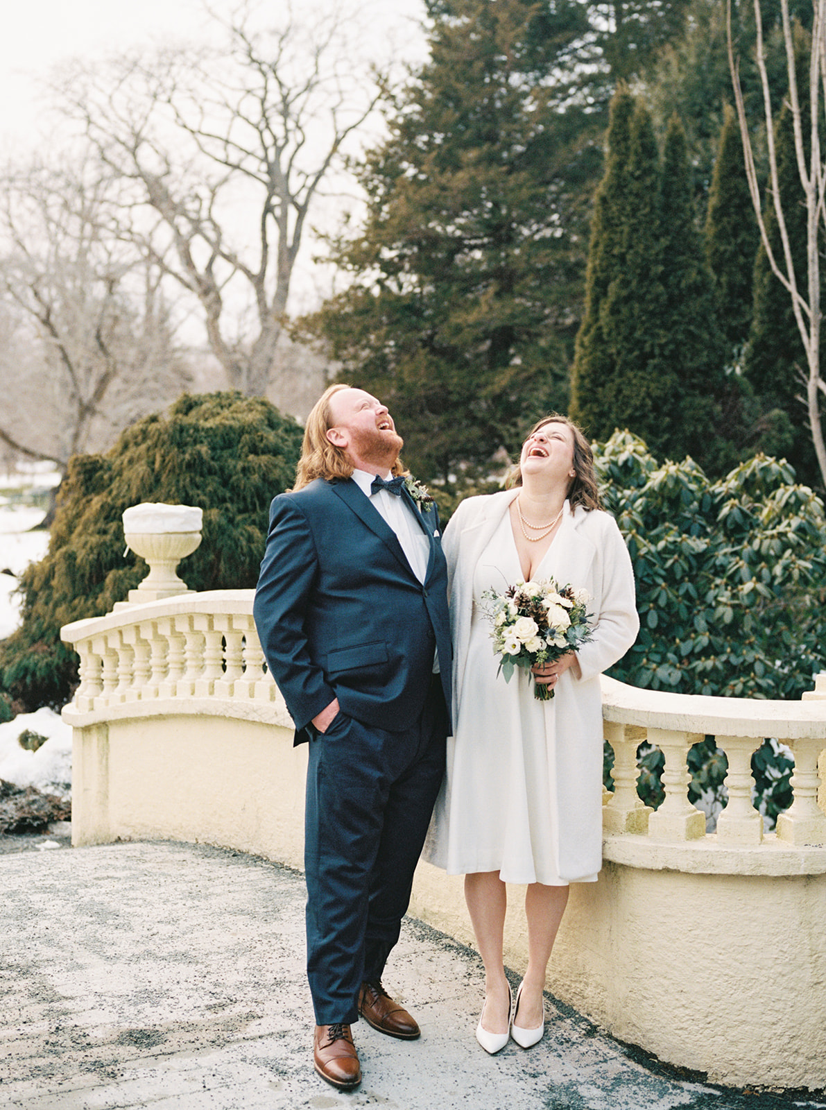 A couple laughs together on an outdoor terrace surrounded by winter greenery and patches of snow, the groom in a navy suit and the bride in a white coat holding a classic bouquet; joyful winter wedding inspiration.