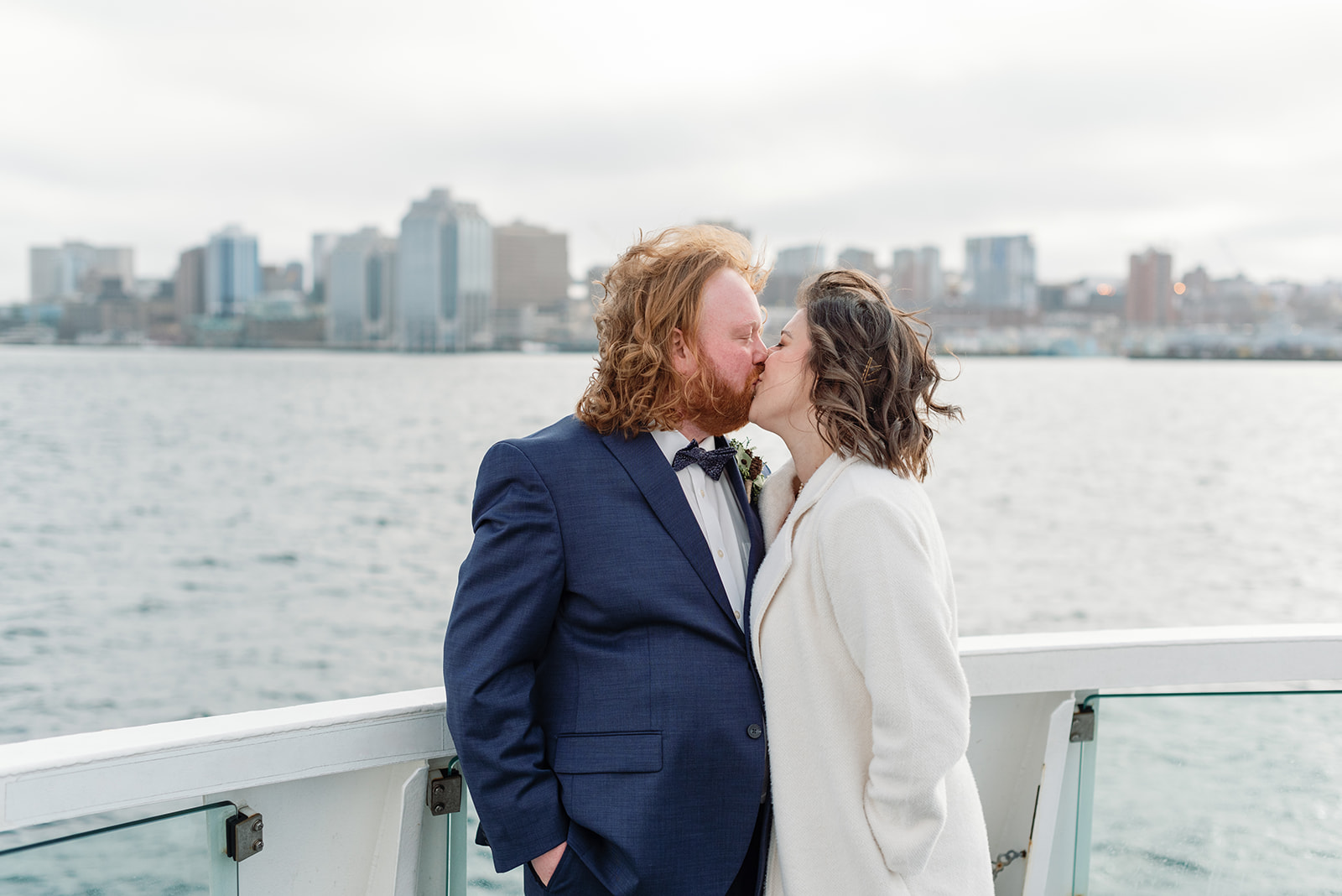 A couple kisses on the deck of a boat with a cold, overcast city skyline behind them; the groom wears a navy suit and the bride a white coat, capturing waterfront winter wedding inspiration.