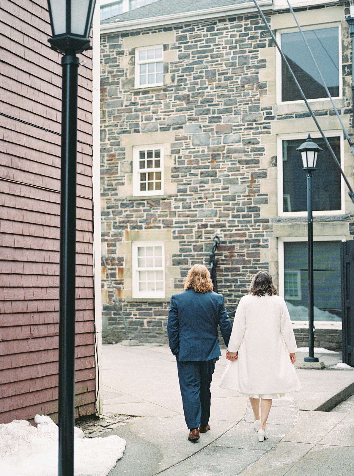 A couple walks hand-in-hand down a historic stone alleyway with patches of snow around them, the groom in a navy suit and the bride in a long white coat.