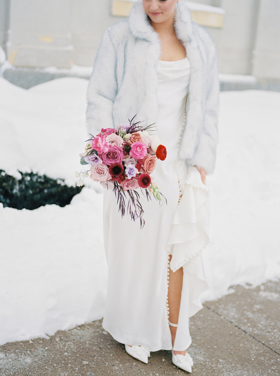 A bride in a pale faux-fur coat and silky white gown holds a bright bouquet of pink, red, and purple flowers, standing in deep snow; soft and romantic winter wedding inspiration.