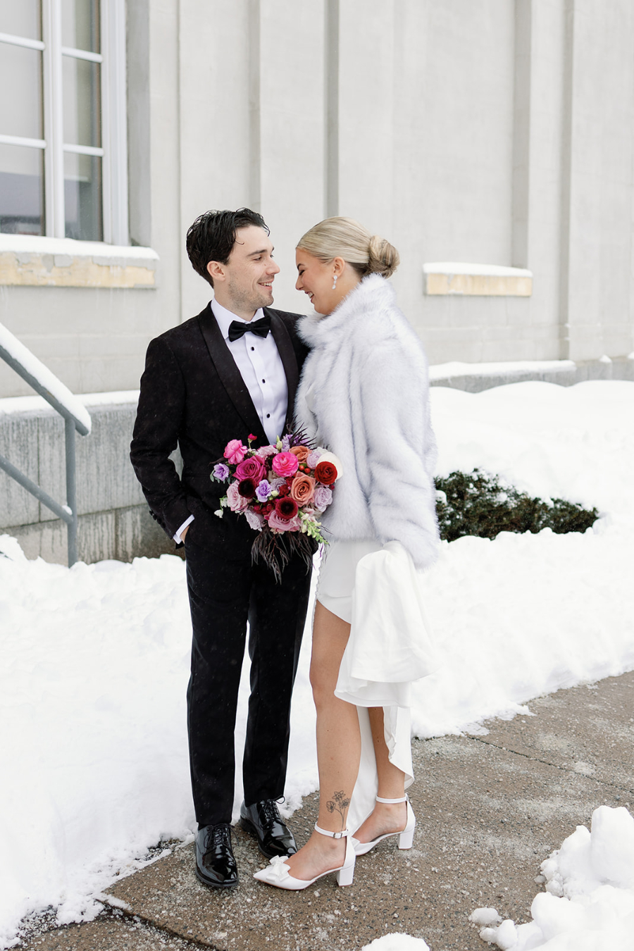 A bride and groom stand close together in fresh snow outside a stone building, the groom in a black tuxedo and the bride in a white faux-fur jacket and short white dress holding a vibrant bouquet of pink and red flowers