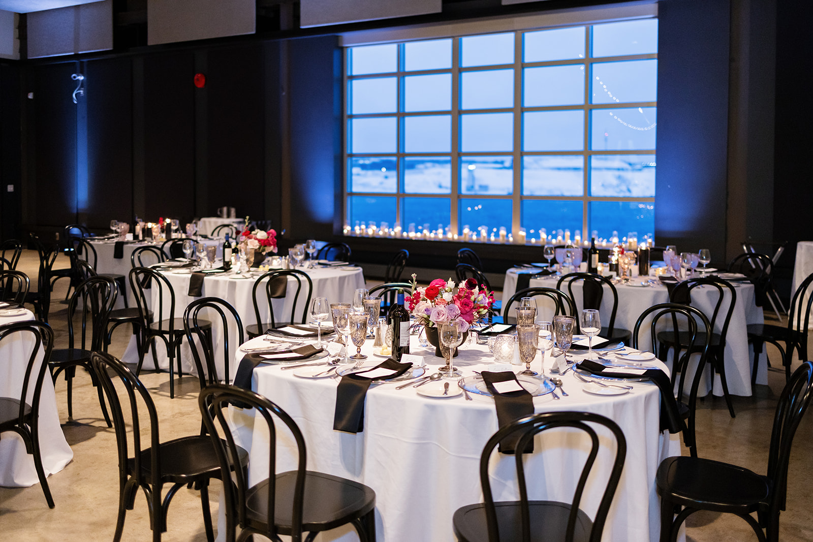 A reception hall set with round tables, white linens, black chairs, and pink floral centerpieces, with icy blue light and snowy views through the windows; elegant indoor winter wedding inspiration.