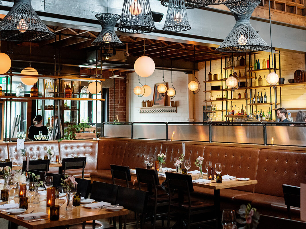 Warm, stylish brasserie with tall woven pendant lights, leather banquettes, set tables with candles and flowers, and an open bar area framed by wood and soft globe lighting.