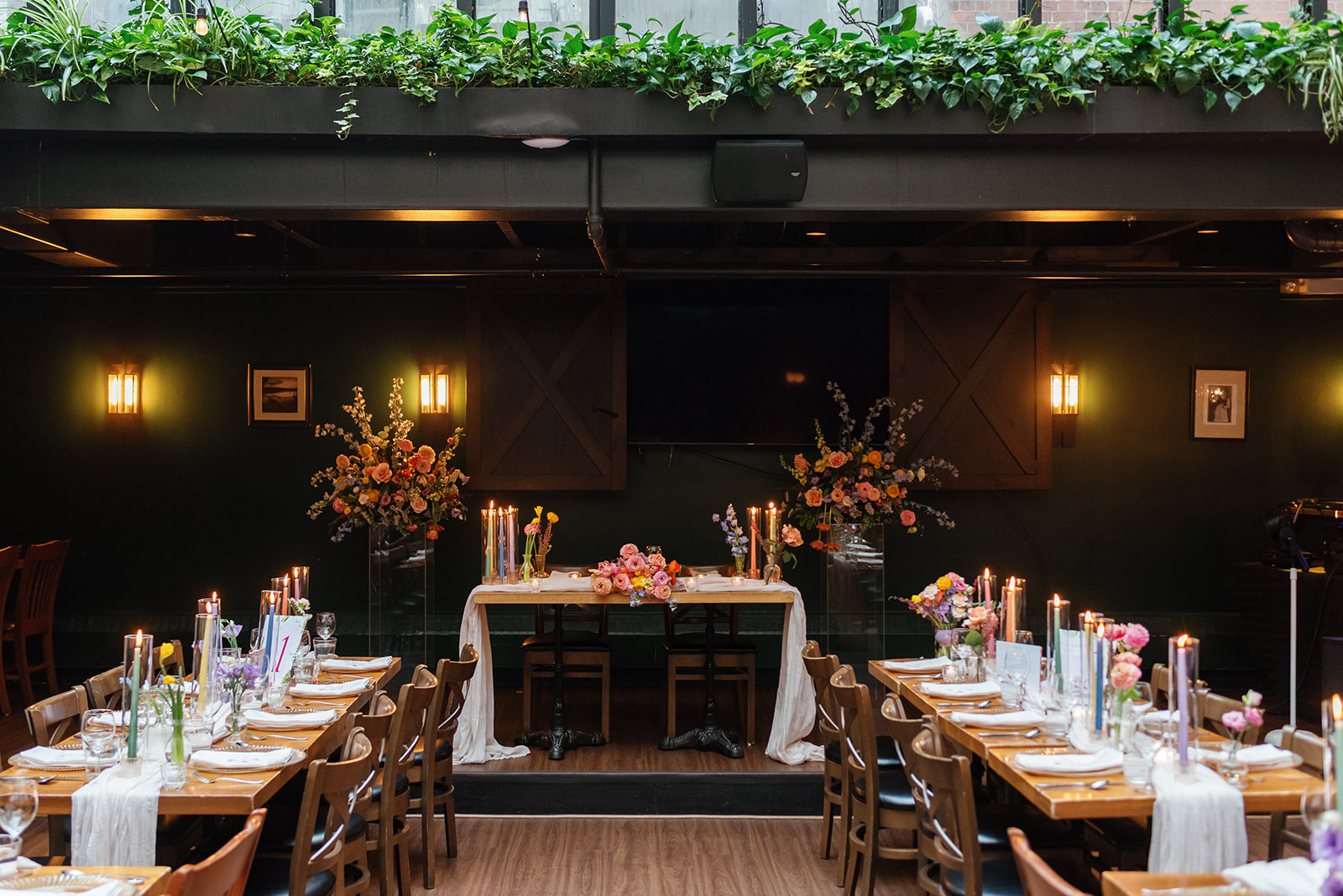 Elegant dining space with long, candlelit tables, lush floral arrangements, dark green walls, warm sconces, and greenery overhead, creating a cozy, intimate atmosphere at Lot Six Bar in Halifax.