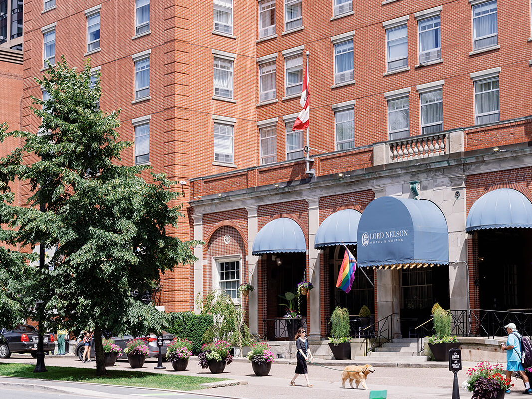Exterior view of the Lord Nelson Hotel & Suites, a historic red-brick building with blue awnings, potted flowers, and flags displayed, including the Canadian and Pride flags; pedestrians walk past on a sunny day.
