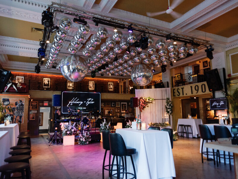 Interior of an event venue featuring a stage framed by floral arrangements and two large screens, with hundreds of disco balls suspended from the ceiling, tall cocktail tables, and dramatic lighting creating a modern celebration space.