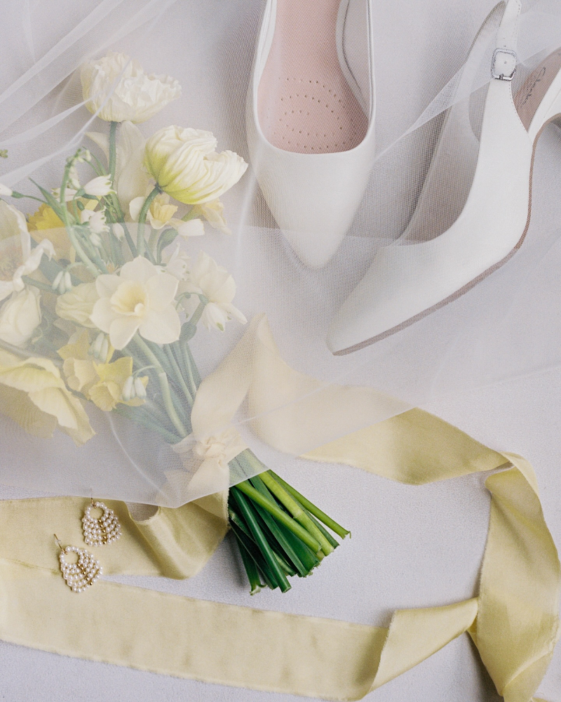 Soft yellow bridal bouquet with silk ribbon and pearl earrings, styled on light fabric with airy, romantic details.